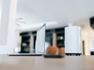 Open laptop on a wooden table beside a white compact desktop/server in a bright open-plan room.